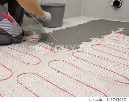 Worker installing electric underfloor heating system mat. Underfloor Heating Installation. Builder applying concrete mortar adhesive with trowel on cables. Energy Efficiency, Smart Home Foundation. Worker installing electric underfloor heating system mat. Underfloor Heating Installation. Builder applying concrete mortar adhesive with trowel on cables. Energy Efficiency, Smart Home Foundation. 141745977