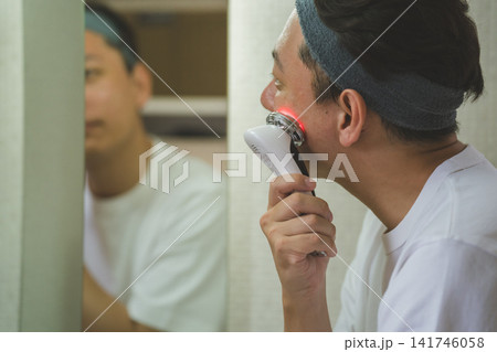 洗面台の鏡の前で多機能美顔器を顎に当てる40代男性のスキンケア 洗面台の鏡の前で多機能美顔器を顎に当てる40代男性のスキンケア 141746058