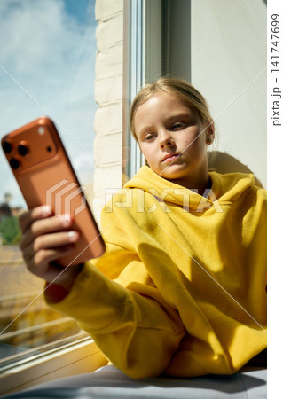 Preteen girl checking smartphone while sitting beside bright window Preteen girl checking smartphone while sitting beside bright window 141747699
