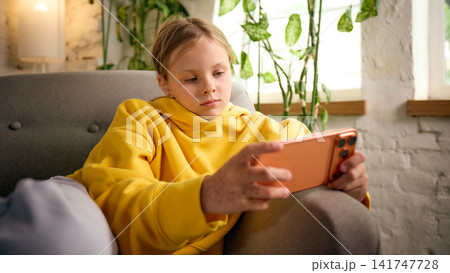 Preteen girl playing mobile game on smartphone while relaxing in armchair 141747728