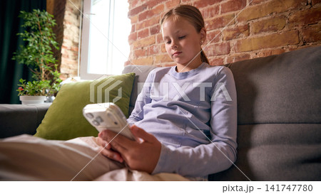 School-age girl scrolling smartphone while relaxing on sofa at home School-age girl scrolling smartphone while relaxing on sofa at home 141747780