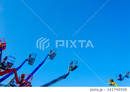 A low-angle, graphic composition capturing a cluster of cherry picker crane arms extending dynamically into a clear blue sky. A low-angle, graphic composition capturing a cluster of cherry picker crane arms extending dynamically into a clear blue sky. 141748509