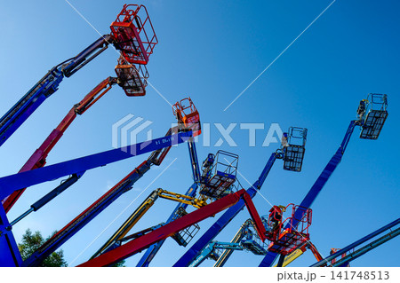 A low-angle, graphic composition capturing a cluster of cherry picker crane arms extending dynamically into a clear blue sky. A low-angle, graphic composition capturing a cluster of cherry picker crane arms extending dynamically into a clear blue sky. 141748513
