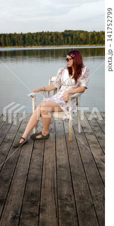 Young woman sits on wooden chair by water at sunset in a dress 141752799