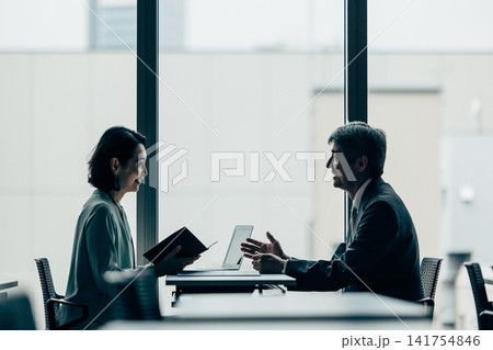 オフィスで対面で会話をする男女ビジネスマンのシルエット 会議イメージ オフィスで対面で会話をする男女ビジネスマンのシルエット 会議イメージ 141754846
