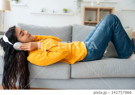 Portrait of Asian woman relaxing listening audio with headphones sitting on a couch at home Portrait of Asian woman relaxing listening audio with headphones sitting on a couch at home 141757866