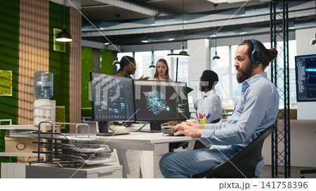 Software developer coding with AI language model assistance in office, listening music. Programmer wearing headphones integrating LLM assistant suggestions into code editor, camera A 141758396