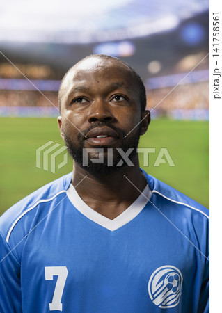 Portrait black soccer player in blue jersey listening to national anthem during pre-match ceremony, match day opening on pitch. Focused footballer standing in lineup. 141758561