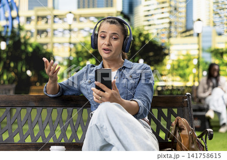Expressive woman wearing over-ear headphones and waving at smartphone screen during video call in city park, showing friendly online greeting and remote interaction. Expressive woman wearing over-ear headphones and waving at smartphone screen during video call in city park, showing friendly online greeting and remote interaction. 141758615