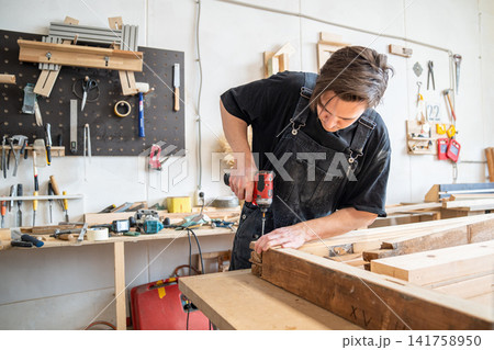 Woodworker technician using handheld cordless electric driver to fasten long screws in wooden frame. Woodworker technician using handheld cordless electric driver to fasten long screws in wooden frame. 141758950