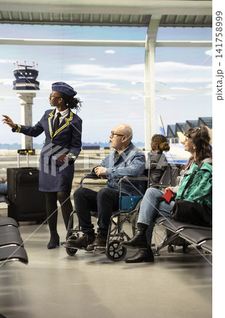 Black flight attendant helping man wheelchair user at terminal gate, providing information about boarding time and giving directions. Old couple receive inclusion, disability-friendly travel. Black flight attendant helping man wheelchair user at terminal gate, providing information about boarding time and giving directions. Old couple receive inclusion, disability-friendly travel. 141758999