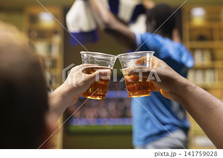 Close up of football supporters drinking beers together while watching match on TV. Back view of fans enjoying alcohol drinks in living room during live game broadcast Close up of football supporters drinking beers together while watching match on TV. Back view of fans enjoying alcohol drinks in living room during live game broadcast 141759028