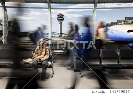 Girl sitting on bench in airport international transport hub surrounded by blurry silhouettes of people walking. Teenage woman waits for plane arrival and crowd rushing in flight departure terminal 141759037