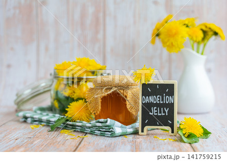 Transparent glass of jar DANDELION JELLY text with dandelion flowers in vase. Soft natural lighting highlights the amber color glossy texture and artisanal character of the preserve. Concept of Transparent glass of jar DANDELION JELLY text with dandelion flowers in vase. Soft natural lighting highlights the amber color glossy texture and artisanal character of the preserve. Concept of 141759215