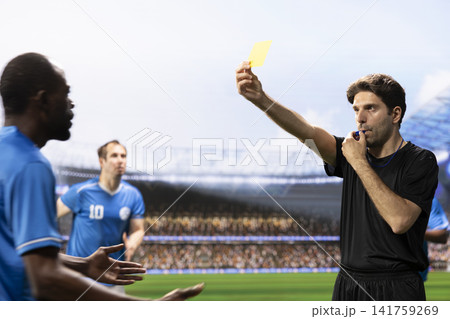 Referee in black uniform showing yellow card and blowing whistle, sanctioning rule violation with card warning. Blue team players protesting referee decision, foul and argument. Referee in black uniform showing yellow card and blowing whistle, sanctioning rule violation with card warning. Blue team players protesting referee decision, foul and argument. 141759269
