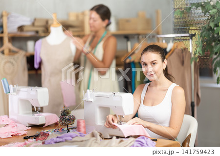 Young tailoress using sewing-machine while other working with mannequin Young tailoress using sewing-machine while other working with mannequin 141759423
