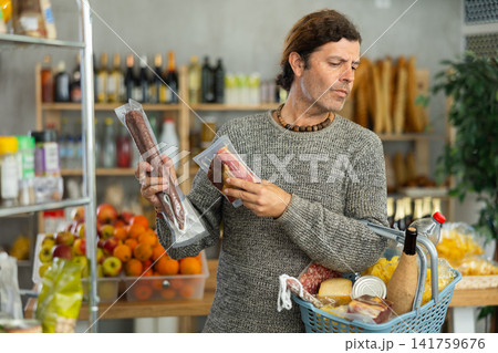 Focused man buyer with shopping basket holding meat sausage at meat department of grocery store 141759676