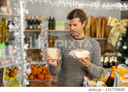 Positive adult man with shopping cart picking out fresh cheese to buy in grocery store for christmas table Positive adult man with shopping cart picking out fresh cheese to buy in grocery store for christmas table 141759680