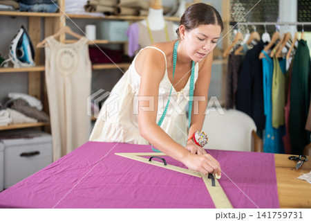 Attentive young female dressmaker drawing on textile in sewing workshop Attentive young female dressmaker drawing on textile in sewing workshop 141759731