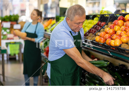 Focused mature salesman laying out small green cucumbers in supermarket Focused mature salesman laying out small green cucumbers in supermarket 141759786