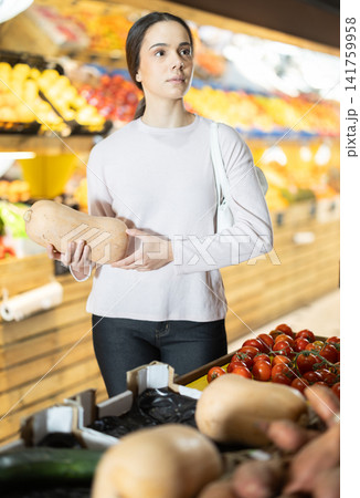 woman stands near a vegetable counter and chooses a ripe pumpkin woman stands near a vegetable counter and chooses a ripe pumpkin 141759958