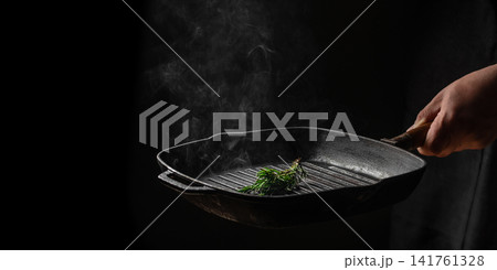 Chef holding hot cast iron grill pan with rosemary and steam on dark background 141761328