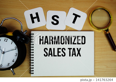 HST Harmonized Sales Tax text written on notepad with magnifying glass and alarm clock on wooden background 141763264