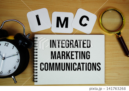 IMC Integrated Marketing Communications text written on notepad with magnifying glass and alarm clock on wooden background 141763268