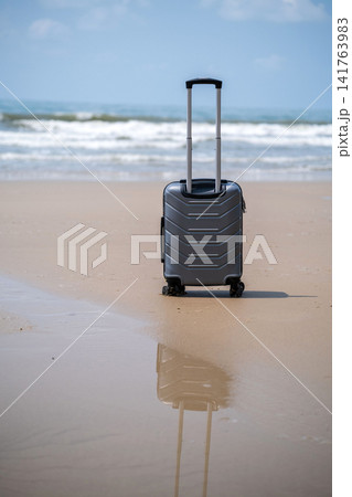 A standalone grey rolling suitcase standing upright on a wide empty sandy beach with calm sea waves and horizon in background. Travel concept A standalone grey rolling suitcase standing upright on a wide empty sandy beach with calm sea waves and horizon in background. Travel concept 141763983
