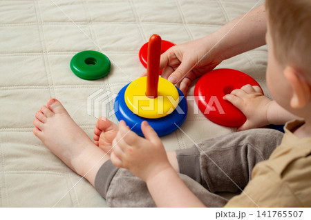 Mother helping child build pyramid on cozy bed. Educational play with shapes and colors. Parent interaction into child game. Family bonding concept Mother helping child build pyramid on cozy bed. Educational play with shapes and colors. Parent interaction into child game. Family bonding concept 141765507
