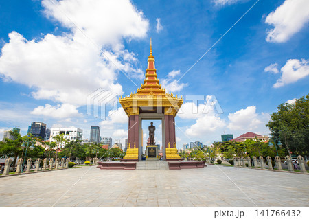 Golden pavilion monument with standing statue in Phnom Penh plaza beneath clear blue sky and city skyline, tropical trees, and ornate roof details Golden pavilion monument with standing statue in Phnom Penh plaza beneath clear blue sky and city skyline, tropical trees, and ornate roof details 141766432