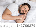 Peaceful sleeping man resting comfortably in a soft bed with white linens. Concept of healthy sleep, wellness, recovery, relaxation, comfort and mental wellbeing. 141766879