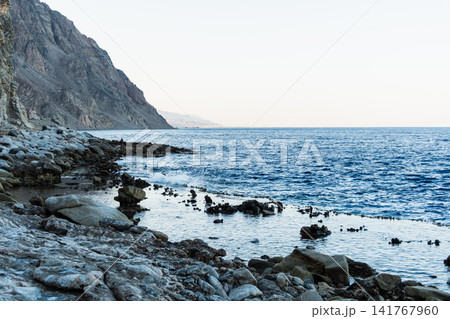 Natural pools with calm blue water along the Red Sea coast in Dahab, Egypt. Rocky shoreline under the Sinai mountains with clear blue water. 141767960