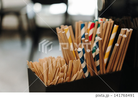 Paper drinking straws and wooden stirrers neatly organized on a coffee shop counter. Eco-friendly accessories for serving beverages Paper drinking straws and wooden stirrers neatly organized on a coffee shop counter. Eco-friendly accessories for serving beverages 141768491