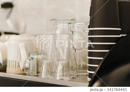 Glass mugs, measuring jiggers and takeaway coffee cups arranged on a professional cafe counter. Equipment for beverage preparation. Glass mugs, measuring jiggers and takeaway coffee cups arranged on a professional cafe counter. Equipment for beverage preparation. 141768493