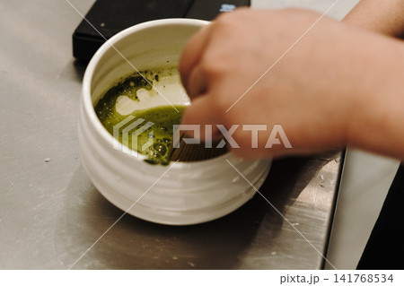 Bamboo whisk mixing matcha tea inside a ceramic bowl to create a smooth texture and foam. Traditional Japanese tea preparation in close-up Bamboo whisk mixing matcha tea inside a ceramic bowl to create a smooth texture and foam. Traditional Japanese tea preparation in close-up 141768534