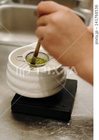 Bamboo whisk gently presses matcha powder through a fine mesh strainer into a ceramic bowl. Traditional tea preparation captured in close-up Bamboo whisk gently presses matcha powder through a fine mesh strainer into a ceramic bowl. Traditional tea preparation captured in close-up 141768536