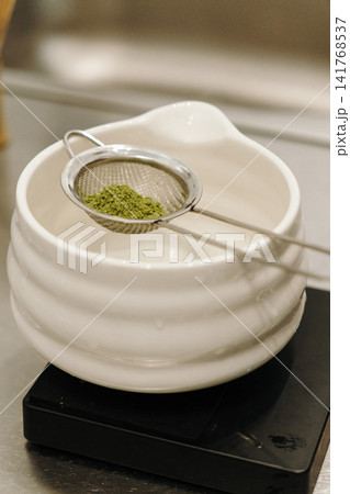 Freshly sifted matcha powder resting in a fine mesh strainer above a ceramic bowl. Traditional tea preparation highlighting premium green tea and brewing equipment Freshly sifted matcha powder resting in a fine mesh strainer above a ceramic bowl. Traditional tea preparation highlighting premium green tea and brewing equipment 141768537