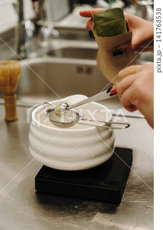 Hands measuring premium matcha powder through a fine mesh strainer into a ceramic bowl. Traditional Japanese tea preparation with precision tools on a digital scale Hands measuring premium matcha powder through a fine mesh strainer into a ceramic bowl. Traditional Japanese tea preparation with precision tools on a digital scale 141768538