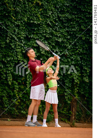 Male coach helping young girl practice tennis serve technique during outdoor lesson Male coach helping young girl practice tennis serve technique during outdoor lesson 141769481