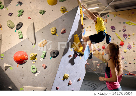 Young girl climbing indoor bouldering wall while female coach watches from below Young girl climbing indoor bouldering wall while female coach watches from below 141769504