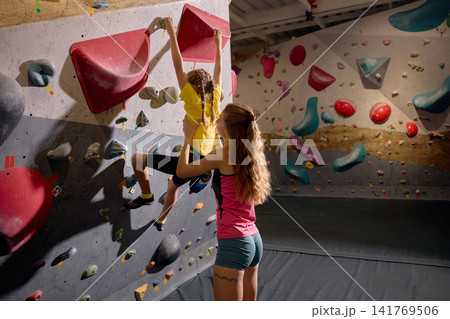 Female coach closely guiding young girl climbing colorful bouldering wall during lesson Female coach closely guiding young girl climbing colorful bouldering wall during lesson 141769506