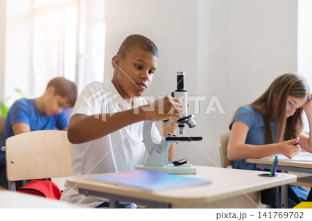 Focused black schoolboy adjusting microscope during practical science lesson in bright classroom 141769702