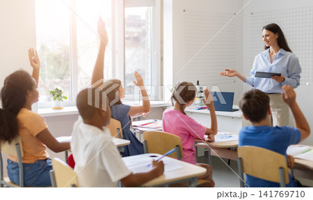 Diverse group of schoolchildren raising hands to answer teachers question during class 141769710