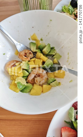 Shrimp and avocado salad with diced mango in a white bowl, two forks resting inside, wooden table surface and additional salad visible in the background Shrimp and avocado salad with diced mango in a white bowl, two forks resting inside, wooden table surface and additional salad visible in the background 141770786