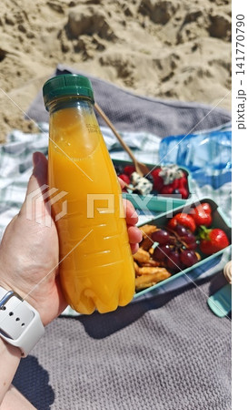 Hand holding a plastic bottle of orange juice near a picnic spread with fruits and snacks on a beach blanket, sand and sunlight visible in the background Hand holding a plastic bottle of orange juice near a picnic spread with fruits and snacks on a beach blanket, sand and sunlight visible in the background 141770790