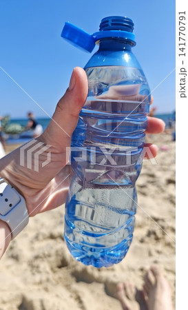 Hand holding a clear plastic water bottle with blue cap against a sunny beach background, sand and people visible in the distance Hand holding a clear plastic water bottle with blue cap against a sunny beach background, sand and people visible in the distance 141770791