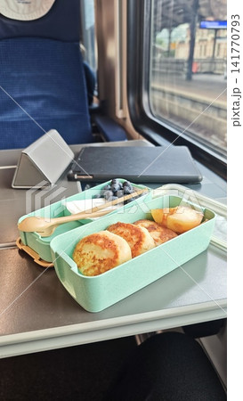 Breakfast meal in a light green container with pancakes, blueberries, and a spoon on a train table, with a blurred view of the station outside the window Breakfast meal in a light green container with pancakes, blueberries, and a spoon on a train table, with a blurred view of the station outside the window 141770793