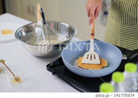 Housewife hand works to make, cook food, expertly flipping golden dessert pancake in kitchen pan, prepared sweet sugar ingredients with whisked batter await more delicious homemade treats 141770874