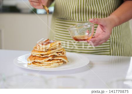 Housewife hands make sweet dessert food, cook sugar ingredients in bright modern kitchen, pouring rich golden syrup on fluffy stack of freshly prepared pancakes, creating delicious meal for morning Housewife hands make sweet dessert food, cook sugar ingredients in bright modern kitchen, pouring rich golden syrup on fluffy stack of freshly prepared pancakes, creating delicious meal for morning 141770875